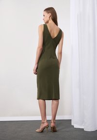 Rochie ajustată de culoare verde măsliniu cu un design în formă de V la spate, până la genunchi, realizată dintr-un material neted. Sandalele aurii cu bretele completează ținuta.