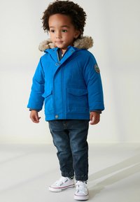 Lysblå oppustelig jakke med falsk pelshætte, to forlommer og et mærke på ærmet. Bæres med mørke denim bukser og hvide sneakers.