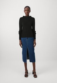 Pull noir côtelé avec un col montant, associé à une jupe en denim bleu arrivant au genou avec une fente devant. Sandales à talons bloc et lanières noires.