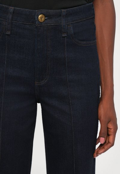 Dark blue denim jeans with front seam stitching, brass button closure, and a hand with painted nails resting at the side.