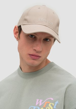 Young man wearing a beige cap with "WRSTBHVR" text and a light grey sweatshirt with colorful "WRST" lettering on chest.