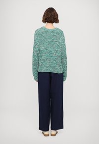 Strickpullover aus türkis- und grün meliertem Garn, mit lockerer Passform, senken Schultern und gerippten Bündchen, kombiniert mit weiten marineblauen Hosen.