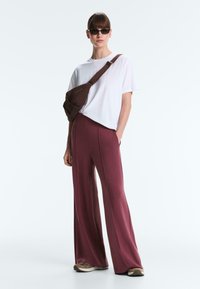 Frau mit Sonnenbrille, weißem T-Shirt, weiten bordeauxroten Hosen, Turnschuhen und einer braunen Umhängetasche, die mit einer Hand in der Tasche steht.