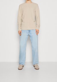 Beige långärmad skjorta med rund halsringning och liten logotyp, matchad med ljusblå raka jeans och beige mocka loafers.
