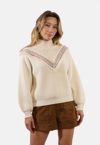 Maglione color crema con collo alto, texture a coste e dettagli in pizzo che formano una forma a V. Maniche lunghe e voluminose con una vestibilità comoda.