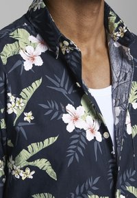 Camicetta blu scuro con un motivo floreale che presenta hibiscus bianchi e fiori gialli, foglie verdi e un colletto con bottoni.