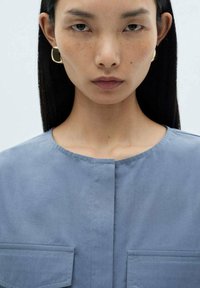 Blue, textured top with a rounded neckline, featuring two front pockets. Model has straight black hair and wears circular gold earrings.