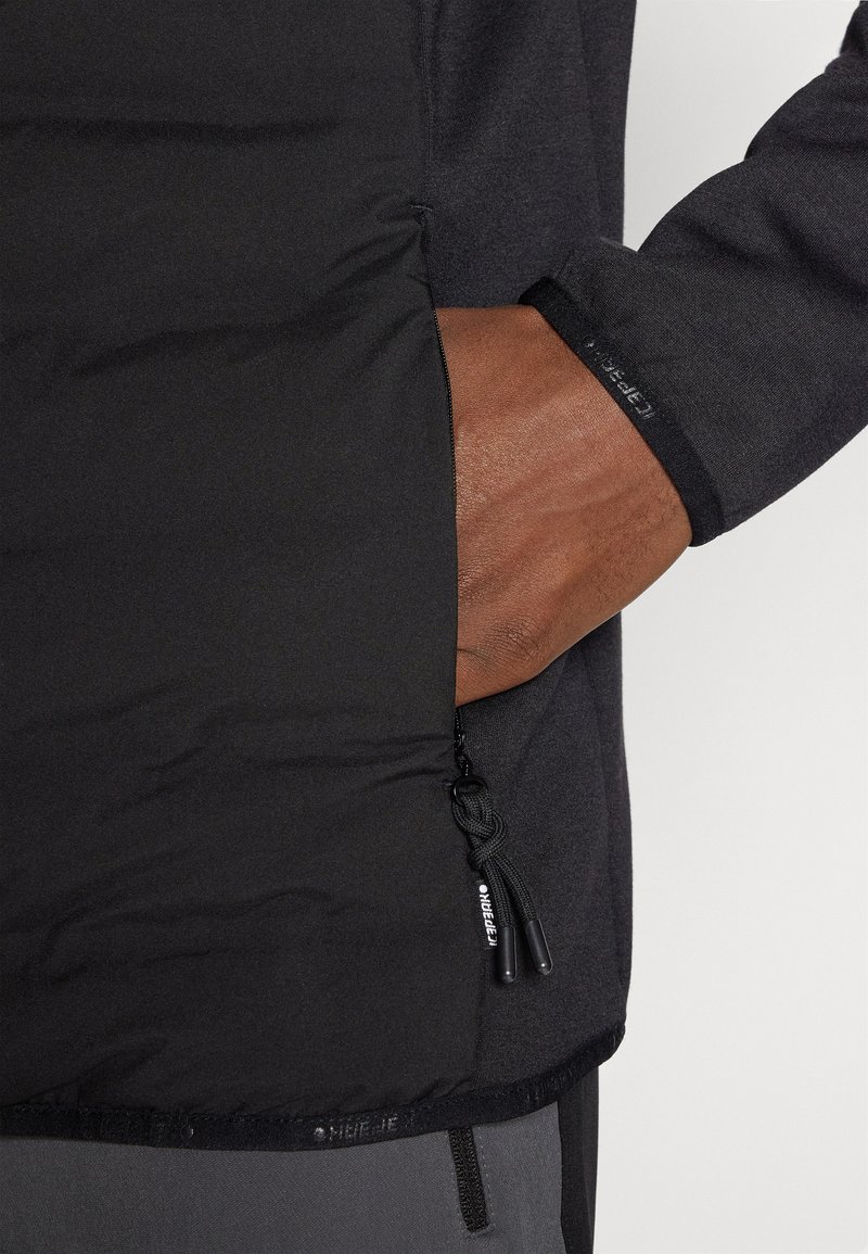 Zwarte geïsoleerde jas met een ritszak, textuurstof en manchetten. Voorzien van trekkoorden en een nauwsluitend ontwerp.