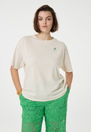 Crème-kleurig t-shirt met een ontspannen pasvorm, korte mouwen en een groen hartlogo. Gecombineerd met groene kanten broek met bladpatronen.