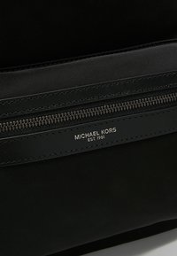 Gros plan sur la section de la fermeture éclair d'un sac Michael Kors noir avec le nom de la marque en argent visible et "EST. 1981" embossé sur une bande en cuir.