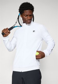 Giacca sportiva bianca con zip, in tessuto testurizzato, dotata di una tasca laterale e un piccolo logo. Indossata con pantaloni scuri e una racchetta da tennis.