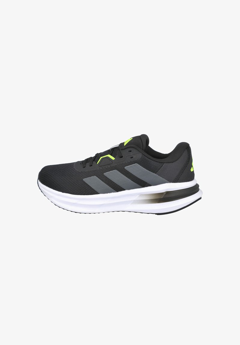 Zapatilla deportiva negra con tejido de malla, acentos grises, detalles en verde brillante y una entresuela blanca; cuenta con un logotipo y una suela texturizada.