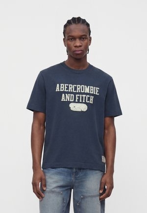 Junger Mann mit geflochtenem Haar, der ein dunkelblaues Abercrombie and Fitch T-Shirt und hellblaue Jeans trägt, steht vor einem einfarbigen weißen Hintergrund.