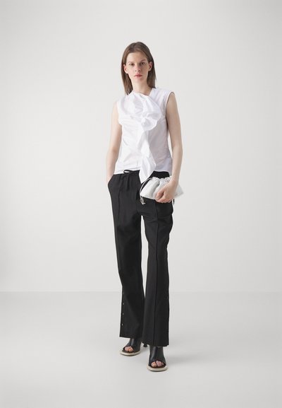 White sleeveless top with ruffles, black pants with button accents, and black sandals. Holding a white, gathered handbag. Minimalistic design.