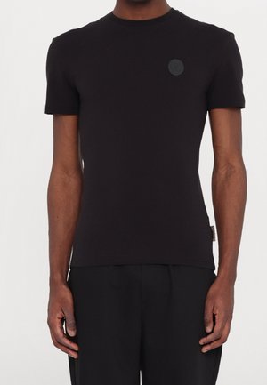 Schwarzes kurzärmeliges T-Shirt aus glattem Stoff. Verfügt über einen runden Ausschnitt und einen kleinen, schwarzen Logopatch auf der linken Brust.