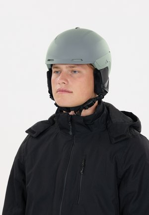 Jonge man met een lichtgrijze helm met zwarte gevoerde oorkappen en een zwarte jas met rits tegen een effen witte achtergrond.