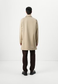 Casaco bege com corte reto e abertura nas costas, combinado com calças castanhas escuras. O tecido parece suave, com um detalhe mínimo.