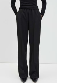 Personne portant un pantalon noir taille haute et large avec les mains dans les poches et des chaussures noires à talons pointus, sur fond léger uni.