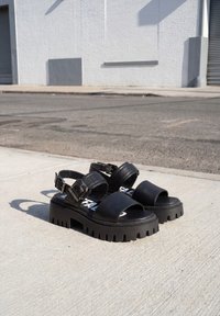 Ζευγάρι μαύρα chunky πλατφόρμα σανδάλια με χοντρές λουριά και μεγάλες αγκράφες τοποθετημένα σε τσιμεντένιο πεζοδρόμιο κοντά σε δρόμο και λευκό τοίχο κτιρίου.