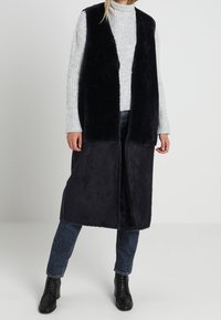 Dlouhá černá vesta s měkkou, kožešinovou texturou, otevřená vpředu a s dvoubarevným designem; sladěná s světle šedým žebrovaným svetrem a džínovými kalhotami.