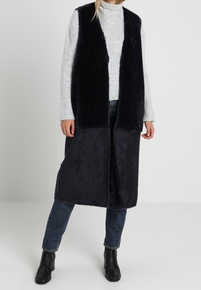 Dlouhá černá vesta s měkkou, kožešinovou texturou, otevřená vpředu a s dvoubarevným designem; sladěná s světle šedým žebrovaným svetrem a džínovými kalhotami.
