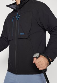 Veste imperméable noire avec des accents de zip bleus, grandes poches avant et un col haut, accompagnée d'un baton de trekking gris.