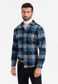 Camisa de franela a cuadros azul y gris con cuello, botones al frente y un bolsillo en el pecho. Se lleva sobre un suéter de cuello alto blanco y se combina con unos jeans oscuros.