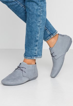 Bottines grises claires à lacets portées avec un jean bleu retroussé sur un fond clair uni.