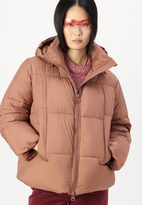 Brun puffjakke med hette, med kviltet tekstur, glidelås foran og store lommer. Bært over en rosa strikkegenser.