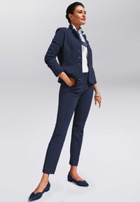 Navy blazer van gestructureerde stof, getailleerd ontwerp, gecombineerd met bijpassende slim fit broek en puntige flats. Witte top en contrasterende patroon sjaal.