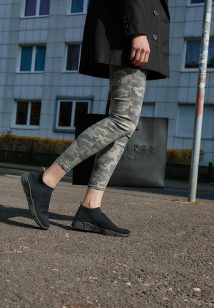 Person går på fortov iført sorte slip-on sneakers, camouflage leggings og en sort frakke, og holder en stor sort indkøbspose.