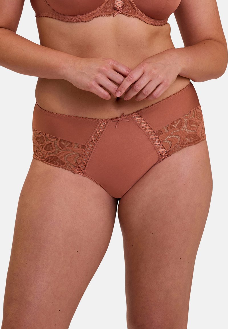 Donna che indossa un reggiseno color ruggine e mutandine abbinate con dettagli in pizzo, mani incrociate in vita, su sfondo chiaro uniforme.