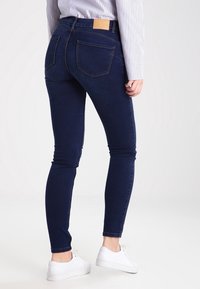 Smala mörkblå jeans med en jämn yta, med två bakfickor och en beige läderlapp vid midjan. Bärs med vita sneakers.