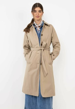 Mujer con gabardina beige con cinturón sobre vestido vaquero azul, con pañuelo estampado en el cuello, manos en los bolsillos y cabello recogido.