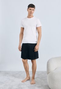 Άσπρο βαμβακερό μπλουζάκι και μαύρο τζιν σορτς με ξεφτισμένα άκρα. Ξυπόλυτος, στέκεται πάνω σε ένα φωτεινό γκρι χαλί σε έναν απλό χώρο.