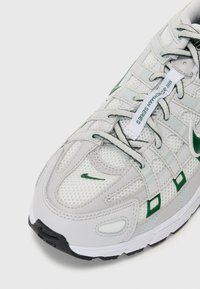 Sneaker in rete grigia con dettagli verde, caratterizzato da un design testurizzato, chiusura con lacci e suola in gomma nera. Logo notevole sul lato.