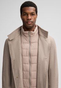 Man in een beige wollen jas over een lichtroze gewatteerde jas, staand voor een effen lichtgrijze achtergrond.