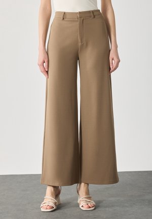 Mujer con pantalones marrones de pierna ancha y cintura alta combinados con sandalias blancas de tacón con tiras, de pie sobre un suelo gris.