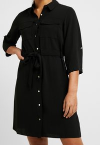 Zwart knopen-overdracht jurk gemaakt van lichtgewicht stof. Voorzien van twee borstzakken, een strikceintuur en opgerolde mouwen met knoopaccenten.