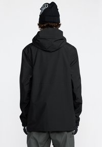 Veste noire imperméable avec capuche, présentant un design minimaliste, des poignets élastiques et une coupe droite, portée sur un pantalon vert.