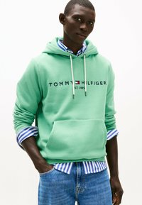 Sudadera verde menta con un bolsillo canguro y cordones, con un logo en el centro. Debajo, se ve una camiseta azul y blanca a rayas.