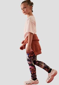 Leggings colorati con un motivo vivace, t-shirt rosa pallido, maglione color ruggine legato in vita e zoccoli bianchi ai piedi.