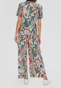 Personne portant un pantalon ample vert assorti et un haut à manches courtes avec un motif floral abstrait coloré, ainsi que des baskets blanches, vue de dos.