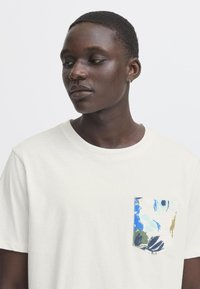 Camiseta de algodón blanca con un bolsillo estampado de flores colorido. Mangas cortas, diseño de cuello redondo, corte holgado, sin herrajes o acentos visibles.