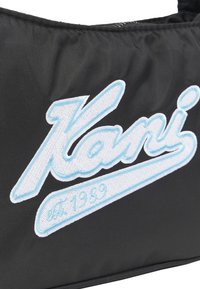 Juodas audinio maišelis su dideliu baltu siuvinėjimu "Kani" su šviesiai mėlynu kontūru ir "est. 1989" mažesniu šriftu žemiau.