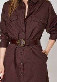 Abito button-up marrone in tessuto leggero, con vita segnato da una cintura con una fibbia in metallo a forma di serpente e due tasche sul petto.