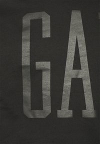 Tela negra con textura mate que presenta letras grandes y audaces en plateado metálico formando la palabra "GAP". El diseño es simple y minimalista.