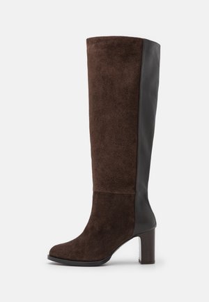 Bota para mujer hasta la rodilla con parte delantera y talón de ante marrón, panel trasero de cuero negro y tacón ancho sobre fondo blanco.