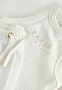 Nahaufnahme eines weißen, gerippten Stofftops mit beigem "BAKER"-Schriftzug und passenden weißen Hosen mit elastischem Bund und Kordelzug.