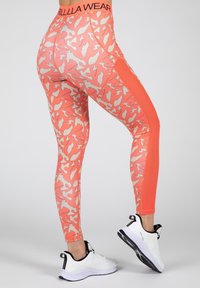 Koraalkleurige leggings met een licht abstract patroon, voorzien van een contrasterend oranje zijpaneel en een zwarte merkband in de taille. Witte sportschoenen.